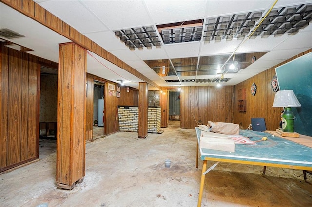 finished basement with visible vents, a paneled ceiling, wooden walls, and water heater