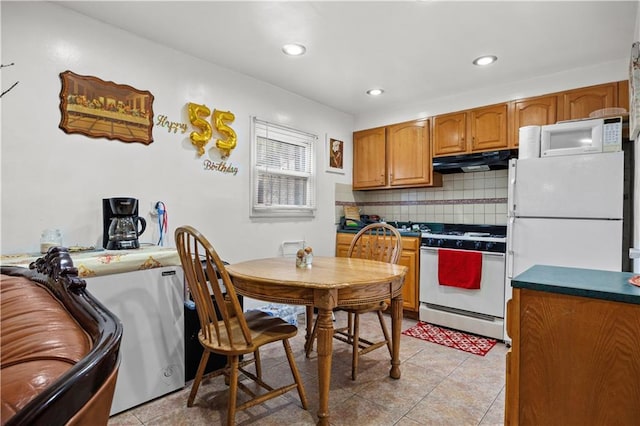 kitchen with under cabinet range hood, white appliances, brown cabinetry, light tile patterned floors, and decorative backsplash