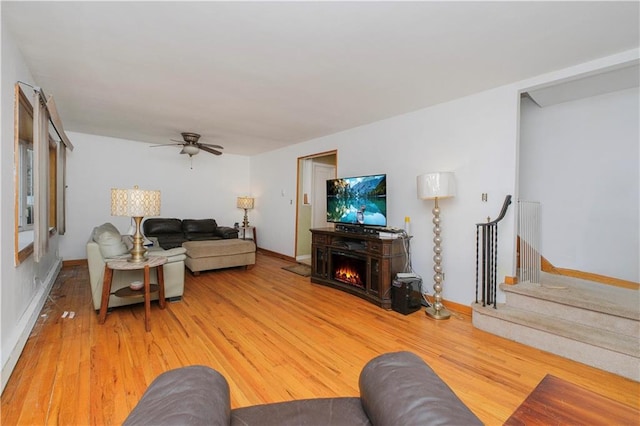 living area with light wood-type flooring, ceiling fan, a warm lit fireplace, baseboards, and stairway
