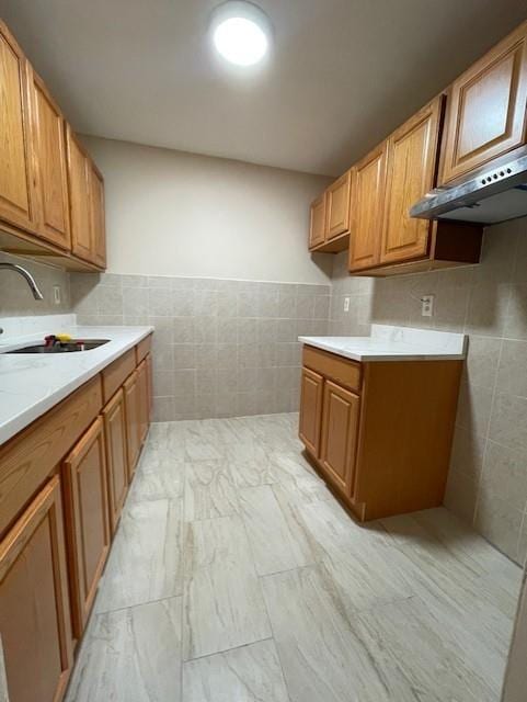 kitchen with tile walls, a sink, under cabinet range hood, light countertops, and brown cabinets
