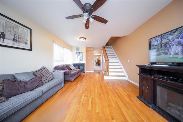living area with light wood-style flooring, ceiling fan, a glass covered fireplace, and stairway