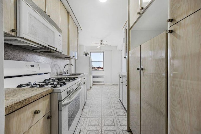 kitchen with a ceiling fan, light brown cabinetry, a sink, radiator heating unit, and white appliances