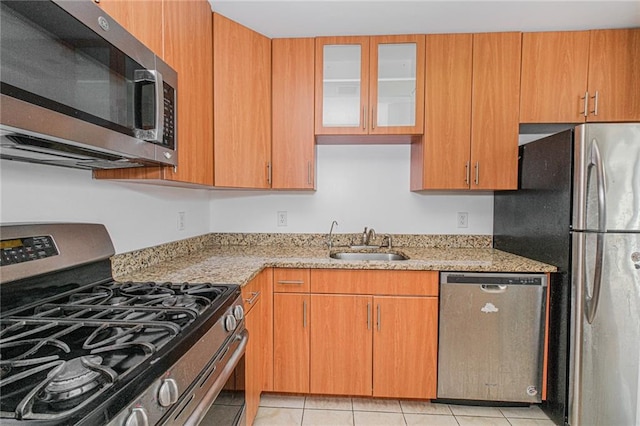 kitchen with appliances with stainless steel finishes, a sink, light stone counters, glass insert cabinets, and light tile patterned flooring