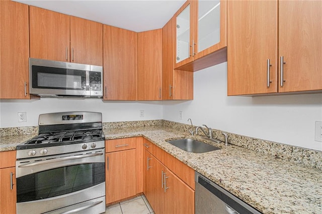 kitchen with appliances with stainless steel finishes, a sink, light stone counters, glass insert cabinets, and light tile patterned floors