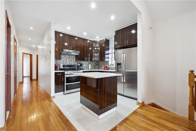 kitchen with stainless steel appliances, light countertops, dark brown cabinetry, under cabinet range hood, and modern cabinets