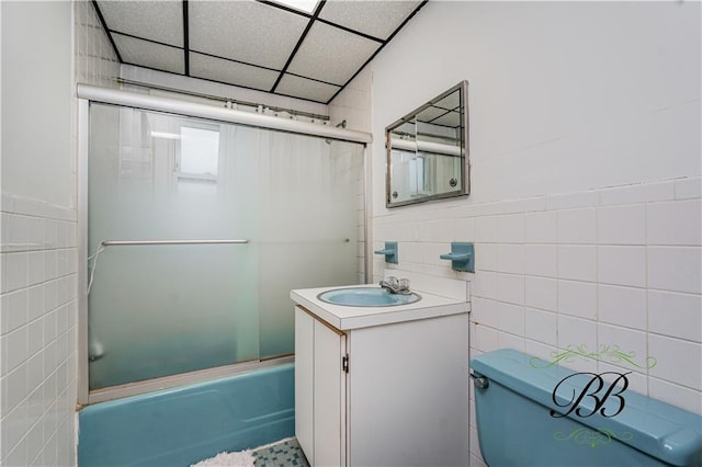 bathroom with vanity, toilet, tile walls, a drop ceiling, and bath / shower combo with glass door