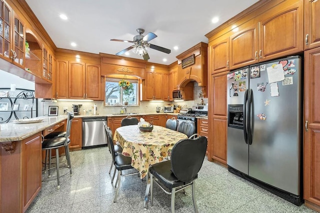 kitchen featuring stainless steel appliances, a ceiling fan, a sink, light stone countertops, and brown cabinetry