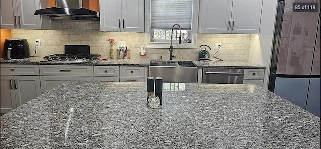 kitchen with wall chimney exhaust hood, appliances with stainless steel finishes, a sink, dark stone counters, and decorative backsplash