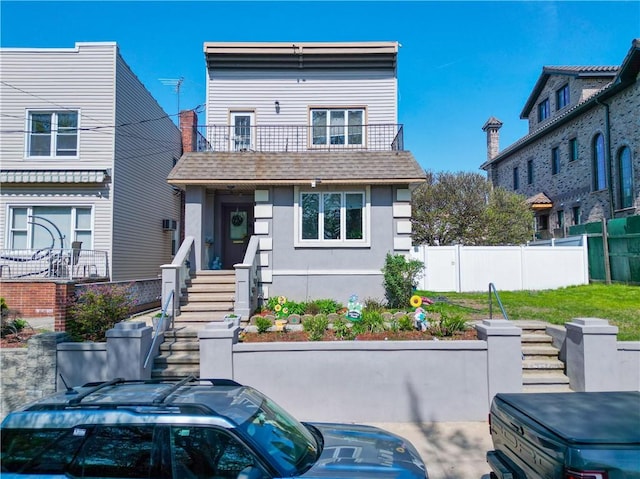 view of front facade featuring fence, a shingled roof, stucco siding, and a balcony