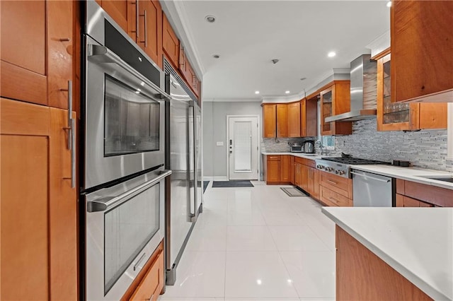 kitchen featuring brown cabinetry, ornamental molding, decorative backsplash, wall chimney range hood, and stainless steel appliances