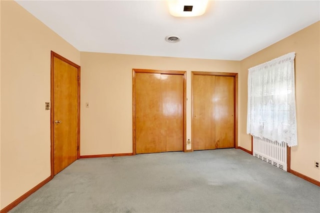 unfurnished bedroom featuring two closets, visible vents, baseboards, light colored carpet, and radiator heating unit