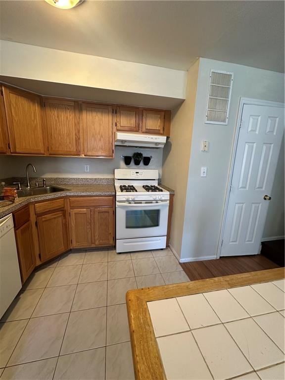 kitchen with white appliances, a sink, under cabinet range hood, light tile patterned floors, and brown cabinetry