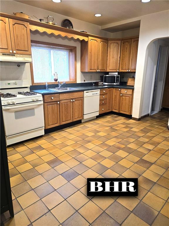 269 Coleridge St, Brooklyn, NY 11235 - kitchen with white appliances, under cabinet range hood, a sink, dark countertops, and arched walkways