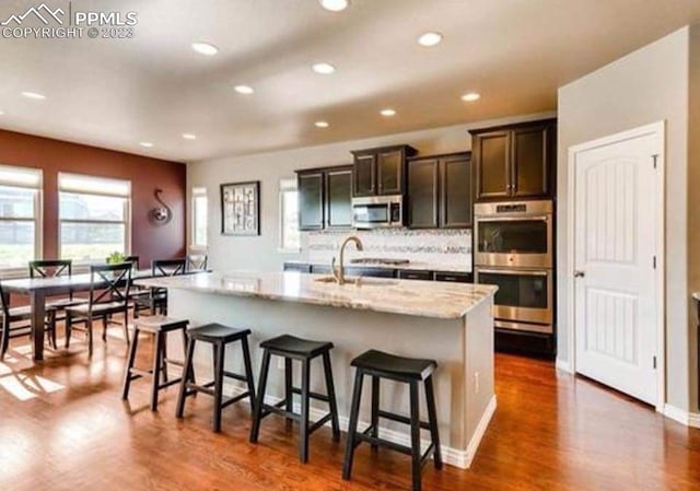 kitchen featuring dark wood-type flooring, tasteful backsplash, an island with sink, light stone countertops, and stainless steel appliances