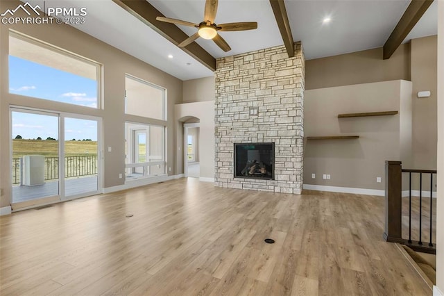 unfurnished living room with a stone fireplace, beamed ceiling, a high ceiling, ceiling fan, and light wood-type flooring