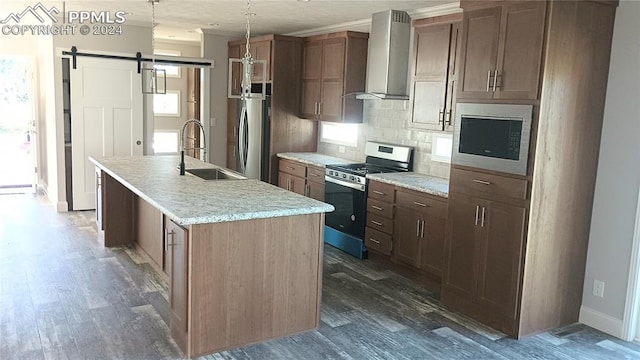 kitchen with wall chimney exhaust hood, tasteful backsplash, a barn door, a kitchen island with sink, and appliances with stainless steel finishes