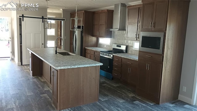 kitchen with backsplash, wall chimney range hood, a barn door, an island with sink, and stainless steel appliances