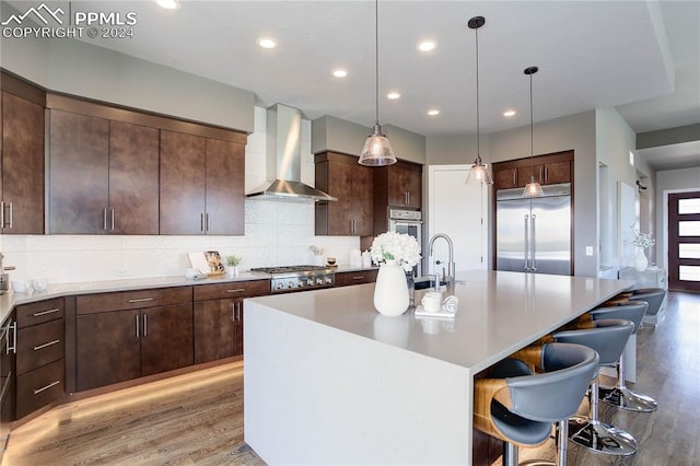 kitchen with wall chimney range hood, hardwood / wood-style flooring, tasteful backsplash, an island with sink, and appliances with stainless steel finishes