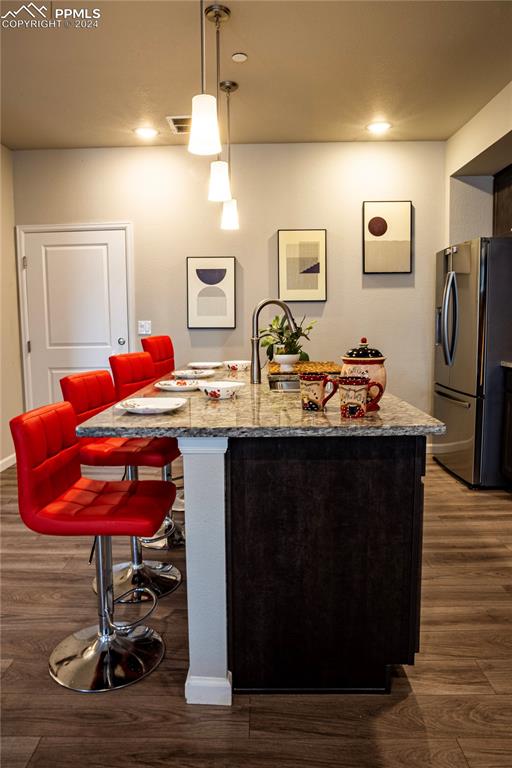 kitchen with a center island with sink, stone counters, stainless steel refrigerator with ice dispenser, decorative light fixtures, and wood-type flooring