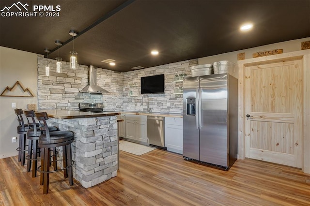 kitchen with wall chimney range hood, light hardwood / wood-style floors, white cabinetry, appliances with stainless steel finishes, and decorative light fixtures