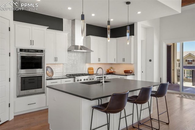 kitchen featuring stainless steel appliances, wall chimney range hood, sink, a center island with sink, and white cabinetry