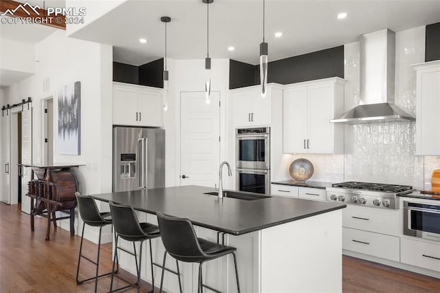 kitchen featuring wall chimney range hood, tasteful backsplash, a barn door, a kitchen island with sink, and appliances with stainless steel finishes