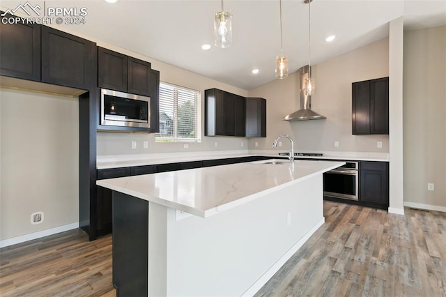 kitchen featuring light hardwood / wood-style floors, a center island with sink, appliances with stainless steel finishes, wall chimney exhaust hood, and pendant lighting