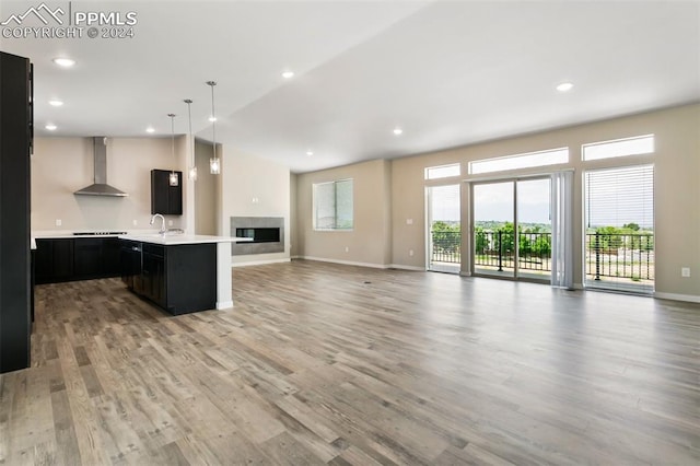 kitchen with wall chimney range hood, a center island with sink, light hardwood / wood-style flooring, decorative light fixtures, and sink