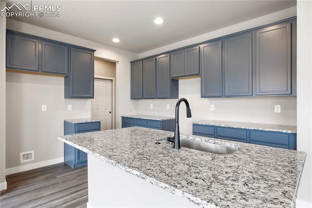 kitchen with wood-type flooring, sink, blue cabinetry, light stone counters, and a kitchen island with sink