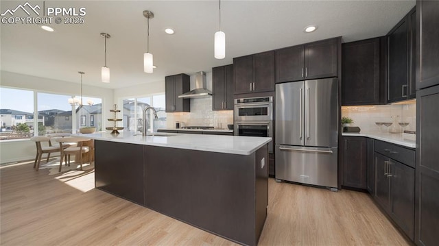 kitchen with sink, decorative light fixtures, appliances with stainless steel finishes, a kitchen island with sink, and wall chimney range hood