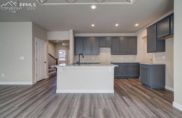 kitchen with light stone counters, a center island with sink, gray cabinets, dark wood-type flooring, and sink