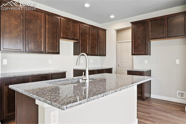 kitchen with wood-type flooring, sink, an island with sink, stone countertops, and dark brown cabinets