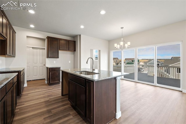 kitchen featuring an inviting chandelier, sink, light stone countertops, an island with sink, and dark brown cabinetry