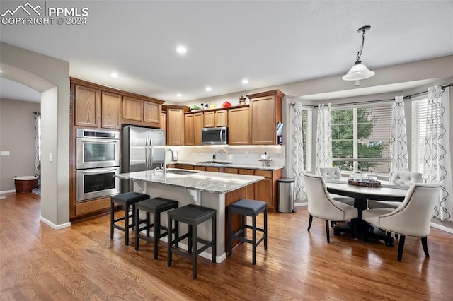 kitchen with light hardwood / wood-style flooring, sink, an island with sink, hanging light fixtures, and appliances with stainless steel finishes