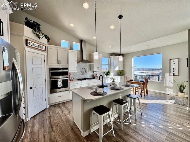 kitchen with white cabinets, hanging light fixtures, a kitchen island with sink, stainless steel appliances, and wall chimney exhaust hood