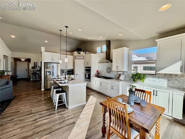 kitchen featuring stainless steel refrigerator with ice dispenser, lofted ceiling, decorative light fixtures, a center island with sink, and wall chimney range hood