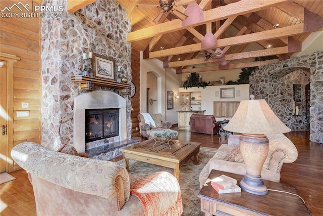 living area with arched walkways, ceiling fan, a stone fireplace, wood finished floors, and beamed ceiling