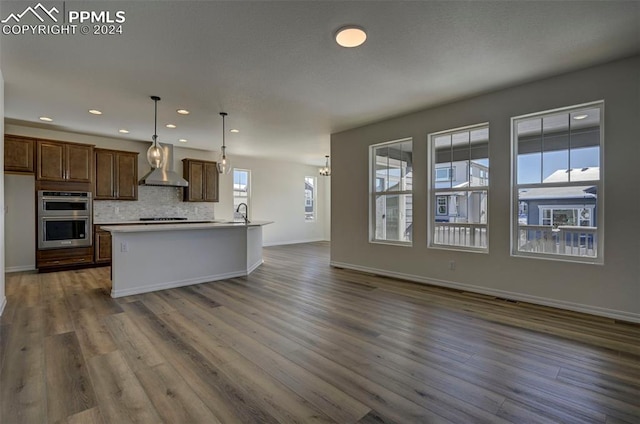 kitchen featuring pendant lighting, a center island with sink, wall chimney exhaust hood, dark hardwood / wood-style floors, and double oven