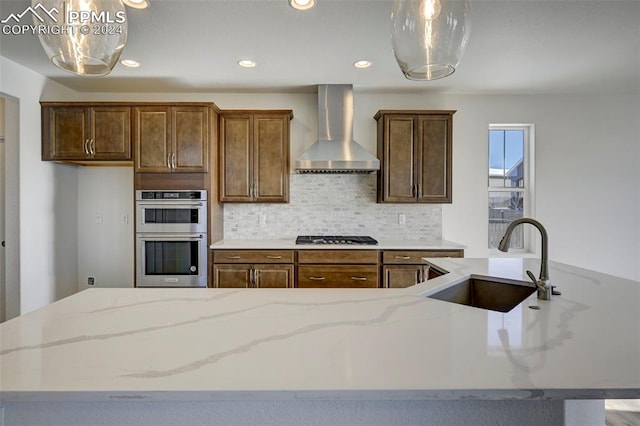 kitchen with light stone countertops, decorative backsplash, appliances with stainless steel finishes, wall chimney exhaust hood, and a sink