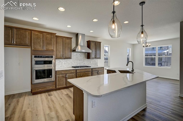 kitchen with a center island with sink, wall chimney range hood, sink, hanging light fixtures, and appliances with stainless steel finishes