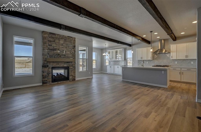 kitchen with backsplash, hanging light fixtures, an island with sink, white cabinets, and wall chimney exhaust hood