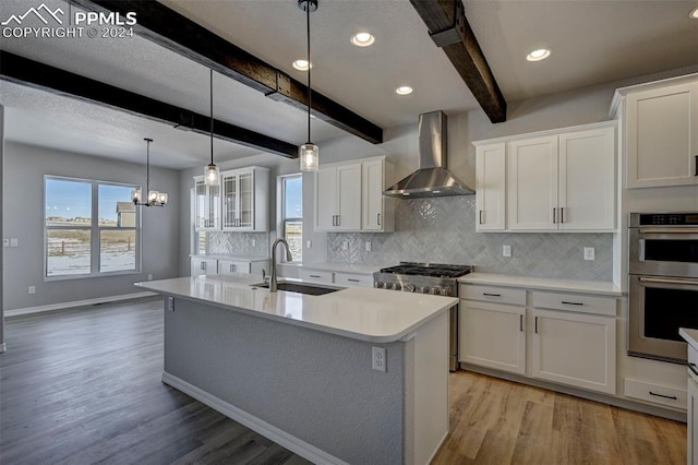 kitchen with sink, double oven, wall chimney range hood, a kitchen island with sink, and white cabinets