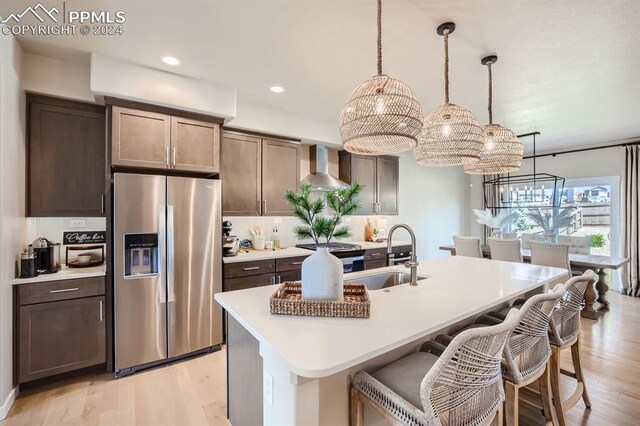 kitchen featuring sink, decorative light fixtures, a kitchen island with sink, wall chimney range hood, and stainless steel refrigerator with ice dispenser