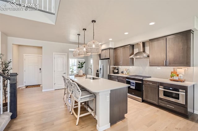 kitchen featuring an island with sink, sink, decorative light fixtures, wall chimney range hood, and appliances with stainless steel finishes