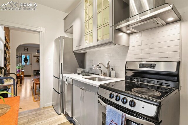 kitchen with gray cabinets, light hardwood / wood-style floors, wall chimney exhaust hood, appliances with stainless steel finishes, and decorative backsplash