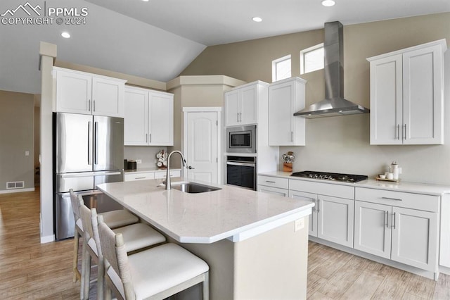 kitchen with wall chimney range hood, sink, white cabinetry, stainless steel appliances, and an island with sink