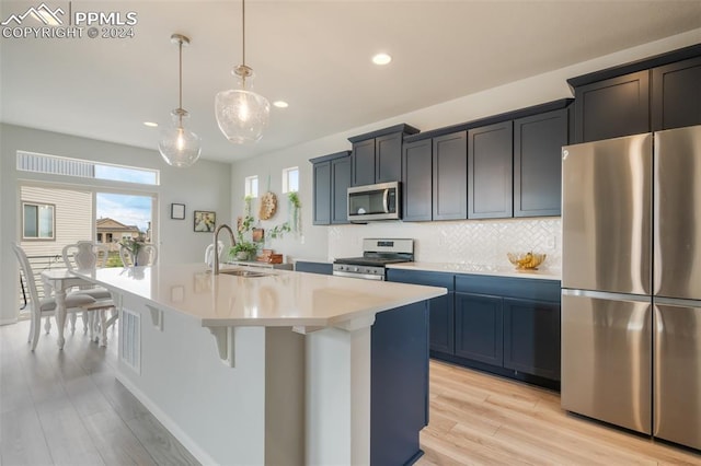 kitchen with hanging light fixtures, stainless steel appliances, light hardwood / wood-style floors, sink, and an island with sink