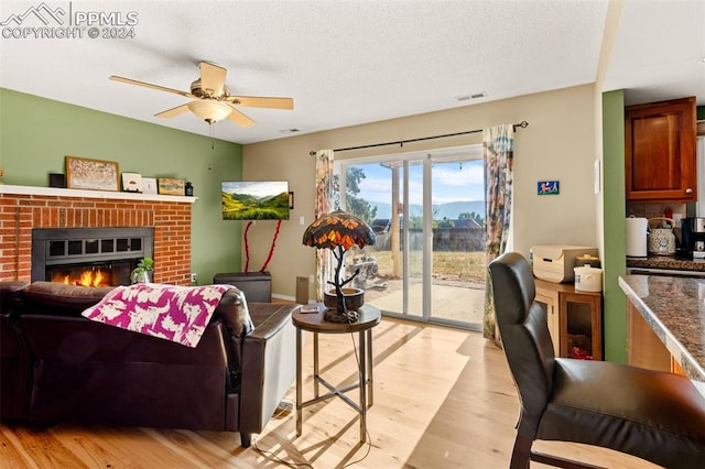 living room featuring ceiling fan, a brick fireplace, a textured ceiling, and light hardwood / wood-style flooring