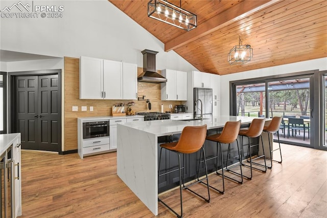 kitchen with a kitchen island with sink, a chandelier, stainless steel appliances, beam ceiling, and wall chimney range hood