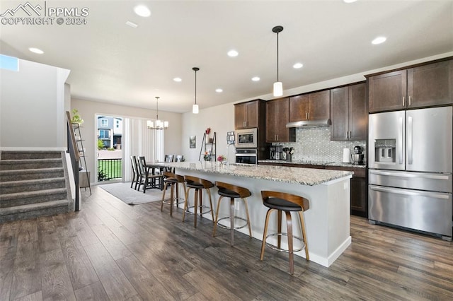 kitchen with dark hardwood / wood-style floors, a notable chandelier, a kitchen island with sink, stainless steel appliances, and light stone counters